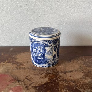 May include: A cylindrical ceramic canister featuring a blue and white transferware pattern. The design includes circular vignettes depicting figures in an interior setting, surrounded by floral motifs. The lid also displays a detailed blue and white scene.