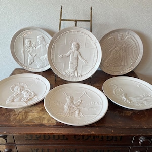 May include: Set of six white ceramic plates with raised images depicting scenes from the Nativity story. The plates are displayed on a wooden shelf with the text "SPODE COTTON" and "WILLIAMANTO" visible.