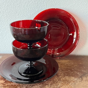 May include: A set of three vintage ruby red glass dessert dishes. The dishes are stacked on top of each other, with the largest dish on the bottom and the smallest dish on top. The dishes have a footed base and a round, shallow bowl.