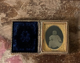 May include: A vintage photograph of a baby in a white dress, framed in a gold-coloured frame with ornate details. The photograph is displayed in a velvet-lined case with a gold-coloured trim.