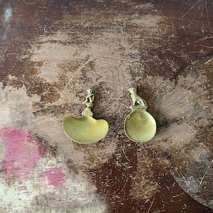 May include: Two small, brass, decorative spoons with a unique design. Each spoon features a small, detailed figure on the handle. The spoons are on a dark brown wooden surface.