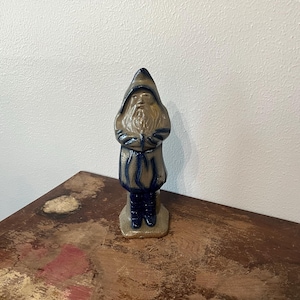 May include: A ceramic figurine of a robed figure with a pointed hood and a long beard. The figure is primarily tan with blue accents on the robe and hood. The figurine is standing on a rectangular base.