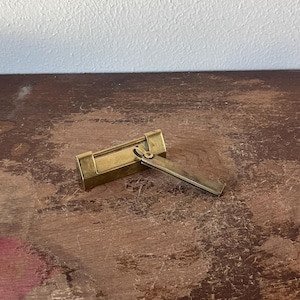 May include: A rectangular brass lock with a hinged, rectangular latch. The lock is open, revealing the interior. The lock and latch are a warm, golden color, set against a distressed, brown wooden surface.
