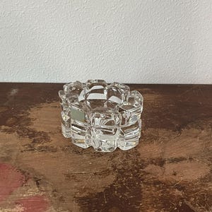 May include: A clear, glass ashtray with a geometric, cube-like design. The ashtray is circular and sits on a wooden surface. The glass is transparent, allowing light to pass through, and the design creates a textured appearance.