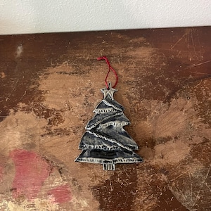 May include: A ceramic Christmas tree ornament with a star on top. The tree is dark gray with white accents and a red string for hanging. The ornament is on a wooden surface.