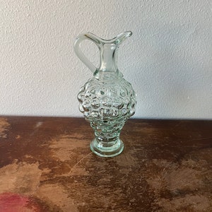 May include: A clear glass bottle with a textured, bubble-like surface. The bottle has a handle and a round base.
