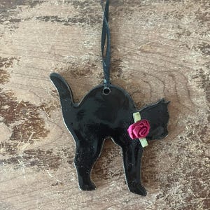 May include: Black cat-shaped ornament with a glossy finish, adorned with a small pink rose and green ribbon. The ornament hangs from a black ribbon. A decorative item for cat lovers.