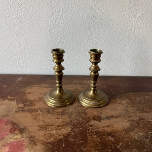 May include: A pair of antique brass candle holders with a simple, elegant design. The holders have a fluted base and a round top.