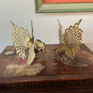 May include: Two gold metal rooster figurines with intricate, openwork wings. The roosters are standing on a wooden surface with the text "SPOOL COTTON WILLIAM ARTIC" painted on it.