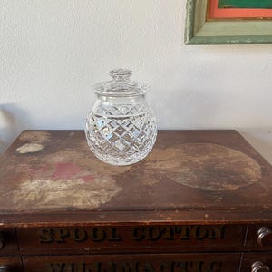 May include: A clear, cut-glass jar with a lid, featuring a diamond pattern. The jar is round and sits on a wooden surface. The words "Spool Cotton" and "Willimantic" are visible on the wooden surface.