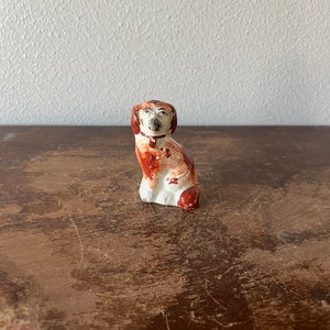 May include: A small, white ceramic figurine of a dog sitting with its head tilted. The dog is painted with brown and red accents.