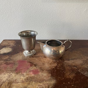 May include: A silver creamer and a silver goblet on a wooden surface with a distressed finish.
