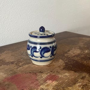 May include: A small, ceramic jar with a lid. The jar is off-white with blue paisley-like designs around the body. The lid has a small knob and a blue rim. The jar is sitting on a wooden surface.