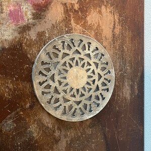May include: A silver-coloured, circular trivet with an intricate, openwork design. The trivet features a central floral motif surrounded by a repeating pattern of stylized shapes. The metal trivet is likely used to protect surfaces from hot cookware.