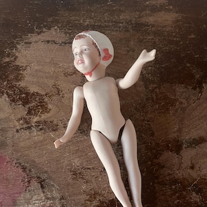 May include: A porcelain doll with a painted face, wearing a white bonnet with red accents. The doll has articulated limbs and is nude, with a light skin tone. The doll is lying on a dark wooden surface.