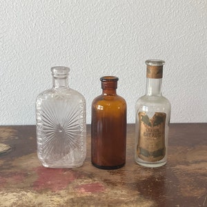 May include: Three glass bottles, one clear with a textured surface, one amber colored, and one clear with a label that reads "Larkin Bay Rug" in gold lettering.