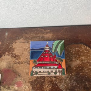 May include: A square ceramic tile featuring a colorful illustration of a building with a red roof, white walls, and a tower. The background shows a blue sky, ocean, and palm trees. The tile is set on a wooden surface.