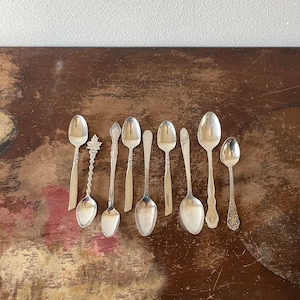 May include: A collection of eight silver spoons with ornate handles, arranged in a row on a dark brown wooden surface.