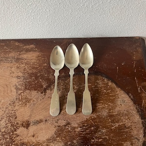 May include: Three silver spoons with oval bowls and elongated handles are arranged on a distressed wooden surface. The spoons have a classic design, reflecting a vintage aesthetic. The spoons are likely for serving or eating.