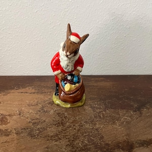 May include: A ceramic figurine of a brown rabbit dressed as Santa Claus, wearing a red suit and hat with white trim. The rabbit is holding a brown sack filled with gold and blue items. The figurine is on a green and brown base.