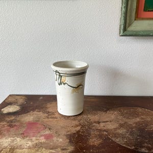 May include: A handmade ceramic tumbler with a cream-colored base. The tumbler features a dark green band near the rim and abstract, hand-painted designs in green and yellow. The tumbler is displayed on a distressed wooden surface.