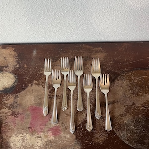 May include: An assortment of eight silver forks with decorative handles, arranged on a dark, distressed wooden surface. The forks have a classic design, with tines and ornate detailing on the handles.