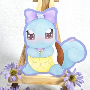 Op de afbeelding: Een lichtblauwe cartoon schildpad sticker met een gele schild, met een klein boeket bloemen. De schildpad heeft een paarse strik en grote, sprankelende ogen. De sticker staat op een klein houten ezel, omringd door paarse bloemen.