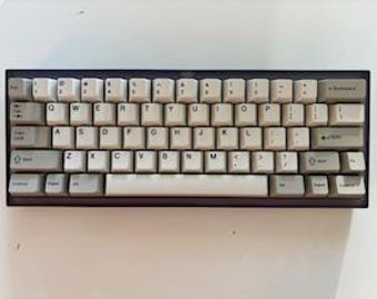May include: A rectangular mechanical keyboard with off-white keycaps and a dark frame. The keyboard has multiple rows of keys, including letters, numbers, and symbols. The keycaps are arranged in a standard QWERTY layout.