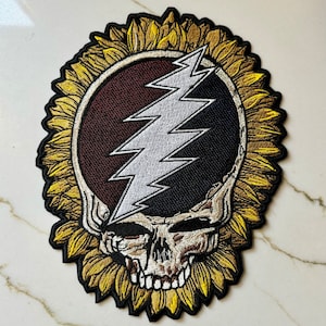 Può includere: Una toppa ricamata in bianco e nero con un design di teschio e fulmine. Il teschio è circondato da un girasole con petali gialli.