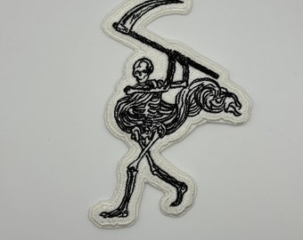 May include: Black and white embroidered patch of a skeleton holding a scythe. The skeleton is walking and has a flowing cape.
