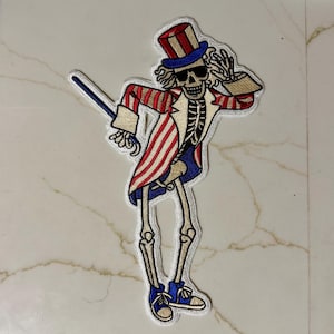 Puede incluir: Parche bordado que representa un esqueleto con un atuendo rojo, blanco y azul al estilo Tío Sam, con sombrero de copa y gafas de sol. El parche tiene un borde blanco y está diseñado para planchar o coser sobre tela.