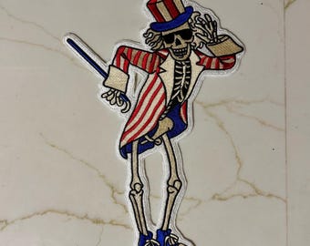 May include: Embroidered patch featuring a skeleton figure in a red, white, and blue Uncle Sam-style outfit, complete with a top hat and sunglasses. The patch has a white border and is designed to be ironed or sewn onto fabric.