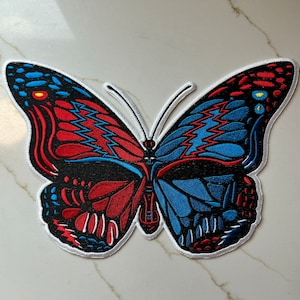Op de afbeelding: Een rode, witte en blauwe vlinderpatch met een bliksemontwerp. De vlinder is geborduurd en heeft een witte omtrek.