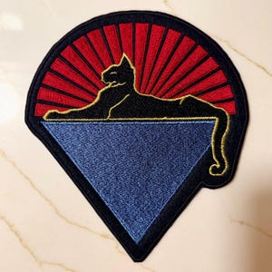Könnte beinhalten: Gestickter Aufnäher mit einer schwarzen Katzen-Silhouette, die sich auf einem blauen Dreieck ausruht. Die Katze ist vor einem roten Sonnenstrahl-Design platziert, alles umrahmt von einem schwarzen Rand. Ein dekorativer Aufnäher.