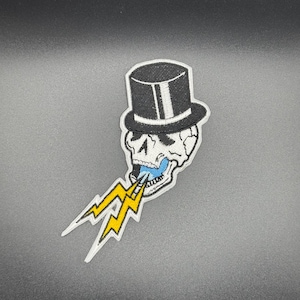 Pode incluir: Um remendo bordado com um crânio usando um chapéu-coco e soprando um raio.