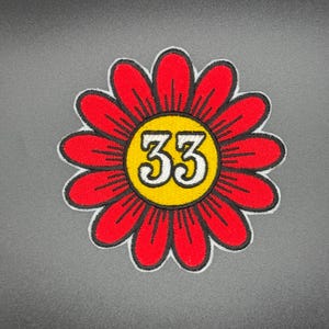 Può includere: Toppa ricamata raffigurante un fiore rosso con centro giallo. Il numero "33" è al centro del fiore, in bianco con contorno nero. La toppa ha un bordo bianco ed è posta su uno sfondo grigio.