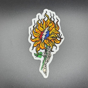 Può includere: Toppa ricamata raffigurante un girasole con un fulmine rosso, bianco e blu al centro. Il girasole è tenuto da una mano scheletrica, con foglie verdi e un bordo bianco. La toppa ha un design artistico unico.