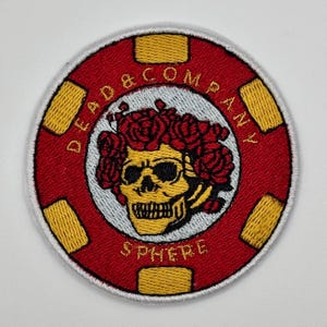 Può includere: Toppa ricamata rossa e gialla con un cerchio bianco al centro. Il cerchio presenta uno scheletro con una corona di rose rosse e il testo "DEAD & COMPANY" sopra e "SPHERE" sotto.