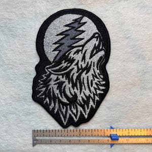 May include: Embroidered patch featuring a howling wolf silhouette against a full moon and a lightning bolt. The patch is outlined in black, with the wolf and moon in shades of gray. The patch measures approximately 4 inches in height.