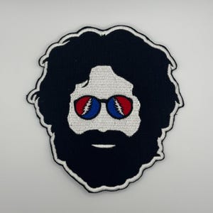 May include: Embroidered patch featuring a stylized face with black hair and beard, and round sunglasses with red and blue accents. The patch has a white border and is likely for clothing or accessories.
