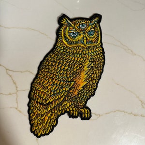 Puede incluir: Un parche de bordado de búho dorado con tres ojos. El búho tiene un diseño detallado con plumas amarillas, naranjas y verdes.