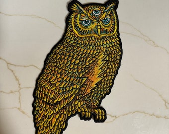 May include: A gold embroidered owl patch with three eyes. The owl has a detailed design with yellow, orange, and green feathers.
