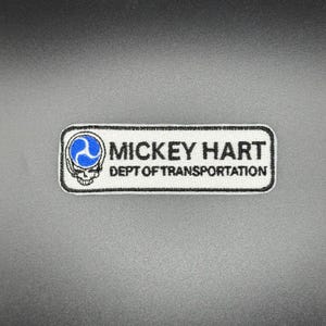 May include: A rectangular patch with a white border and black background. The patch features the text "MICKEY HART DEPT OF TRANSPORTATION" in black. A blue and white skull graphic is on the left side.