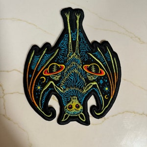 Puede incluir: Un parche bordado negro, azul y amarillo de un murciélago con las alas extendidas. El murciélago tiene un diseño detallado con planetas y estrellas en sus alas.