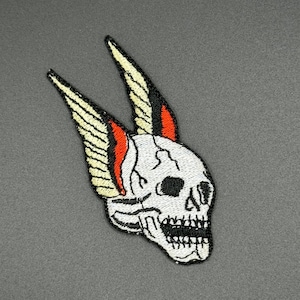 May include: White skull with black eyes and a red and yellow winged design. The skull is embroidered on a grey fabric.