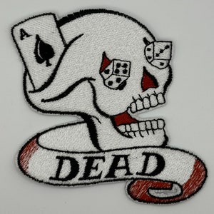 Op de afbeelding: Witte geborduurde patch met een schedel met een enkele speelkaart, de aas van schoppen, en twee dobbelstenen. De schedel heeft een rode mond en het woord "DEAD" staat op een witte banner onder de schedel.