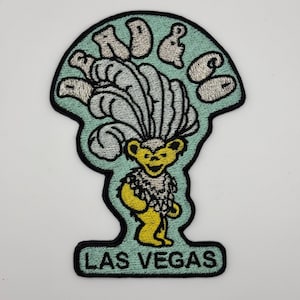 Può includere: Una toppa ricamata blu chiaro con un orso giallo che indossa un copricapo di piume bianche. Il testo "Dead & Co" è sopra l'orso e "Las Vegas" è sotto l'orso.