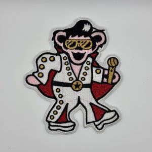 Può includere: Una toppa ricamata con un orso dei cartoni animati vestito come Elvis Presley, con una tuta bianca con dettagli dorati, un mantello rosso e occhiali da sole. L'orso tiene un microfono.