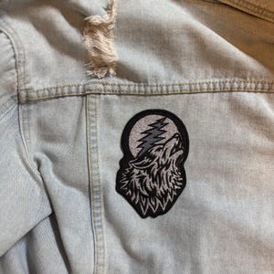 May include: Light blue denim jacket with a distressed upper section. A black and gray embroidered patch of a howling wolf against a moon and lightning design is sewn on the back of the jacket.