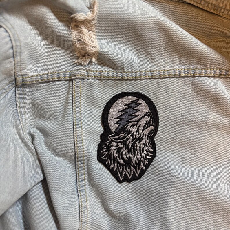 May include: Light blue denim jacket with a distressed upper section. A black and gray embroidered patch of a howling wolf against a moon and lightning design is sewn on the back of the jacket.